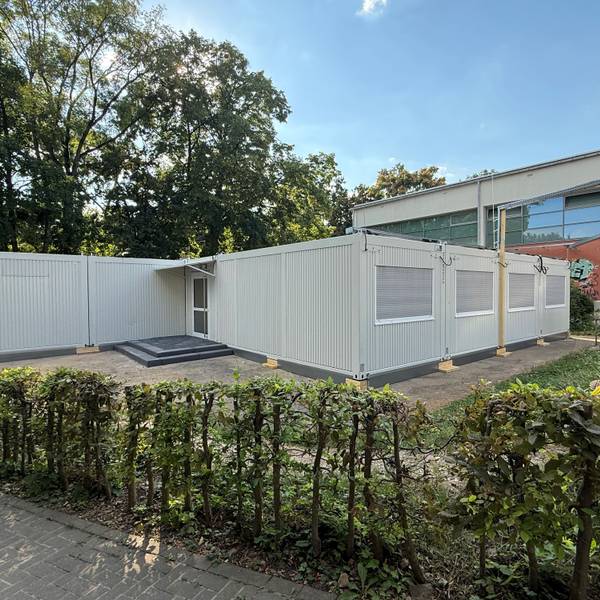 Ein grauer Containerbau steht vor einem Schulgebäude.