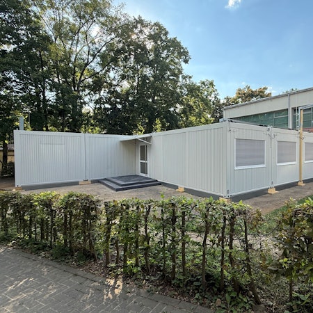 Ein grauer Containerbau steht vor einem Schulgebäude.