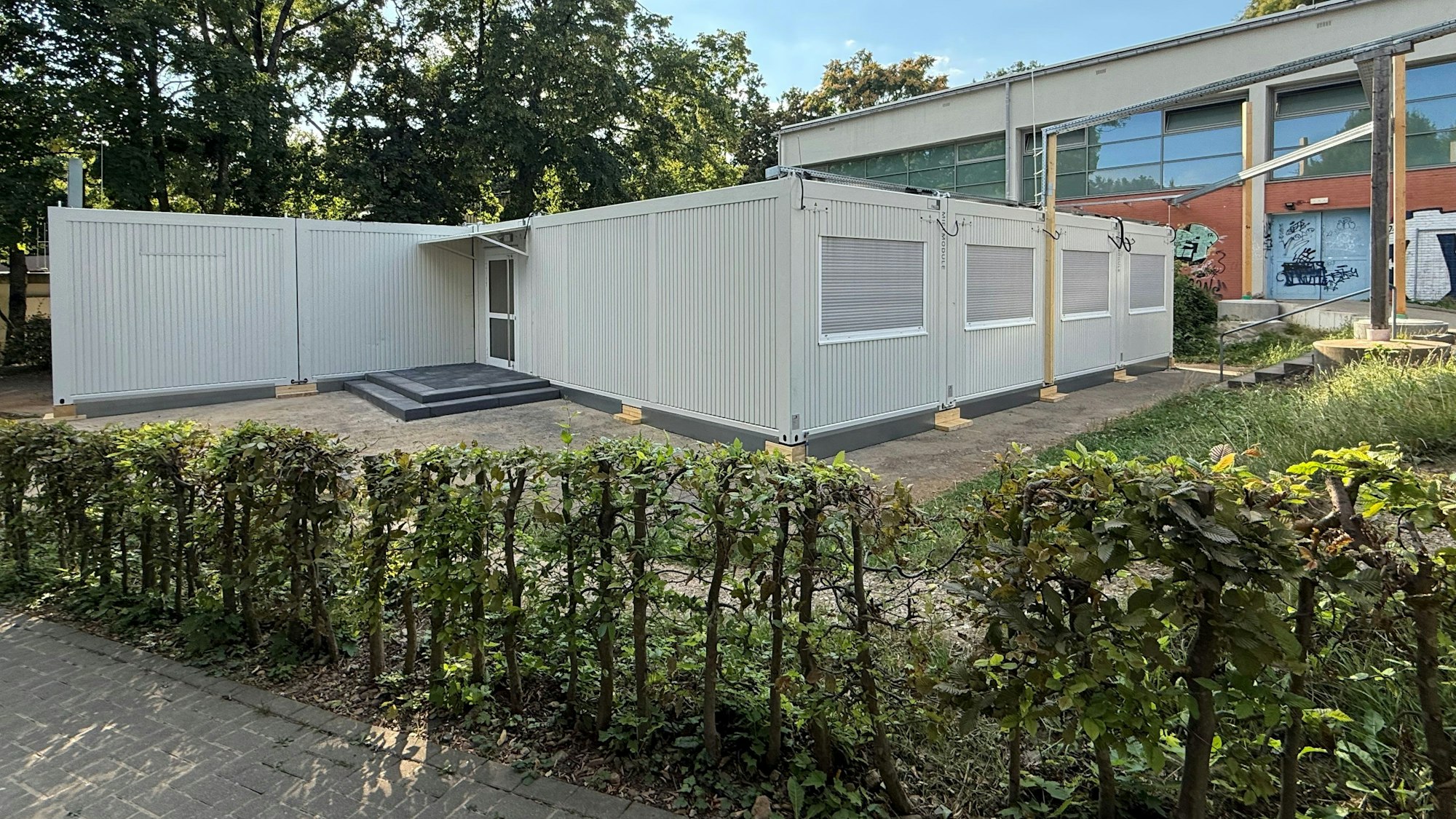 Ein grauer Containerbau steht vor einem Schulgebäude.