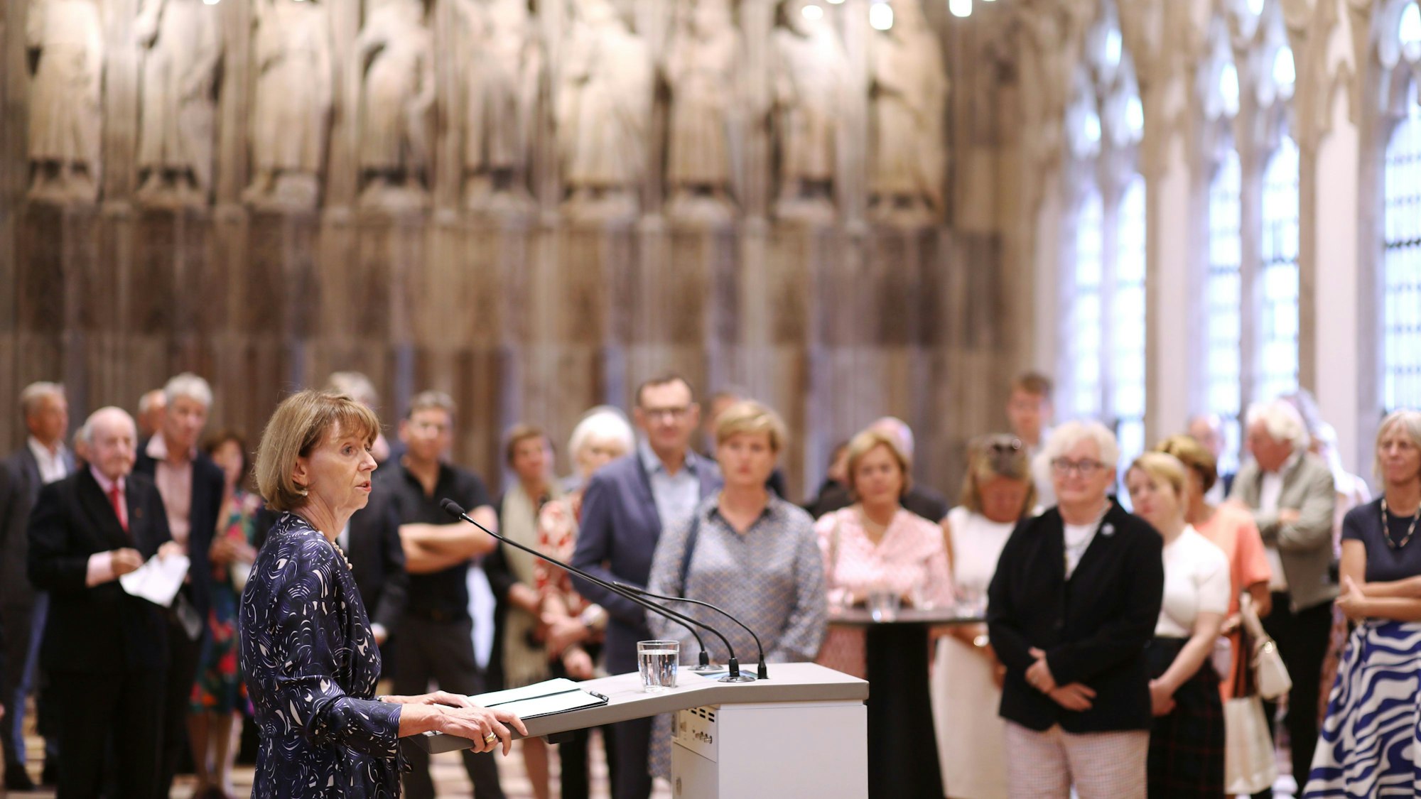 Henriette Reker steht am Rednerpult, im Hintergrund hören Menschen zu.