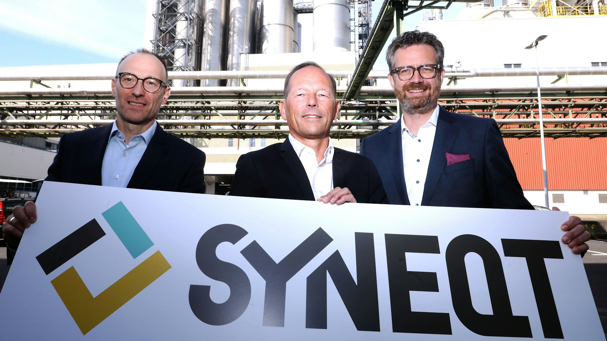 Die GF-Riege ist mit dem Plakat zu sehen, auf dem der Name Syneqt zu lesen ist.