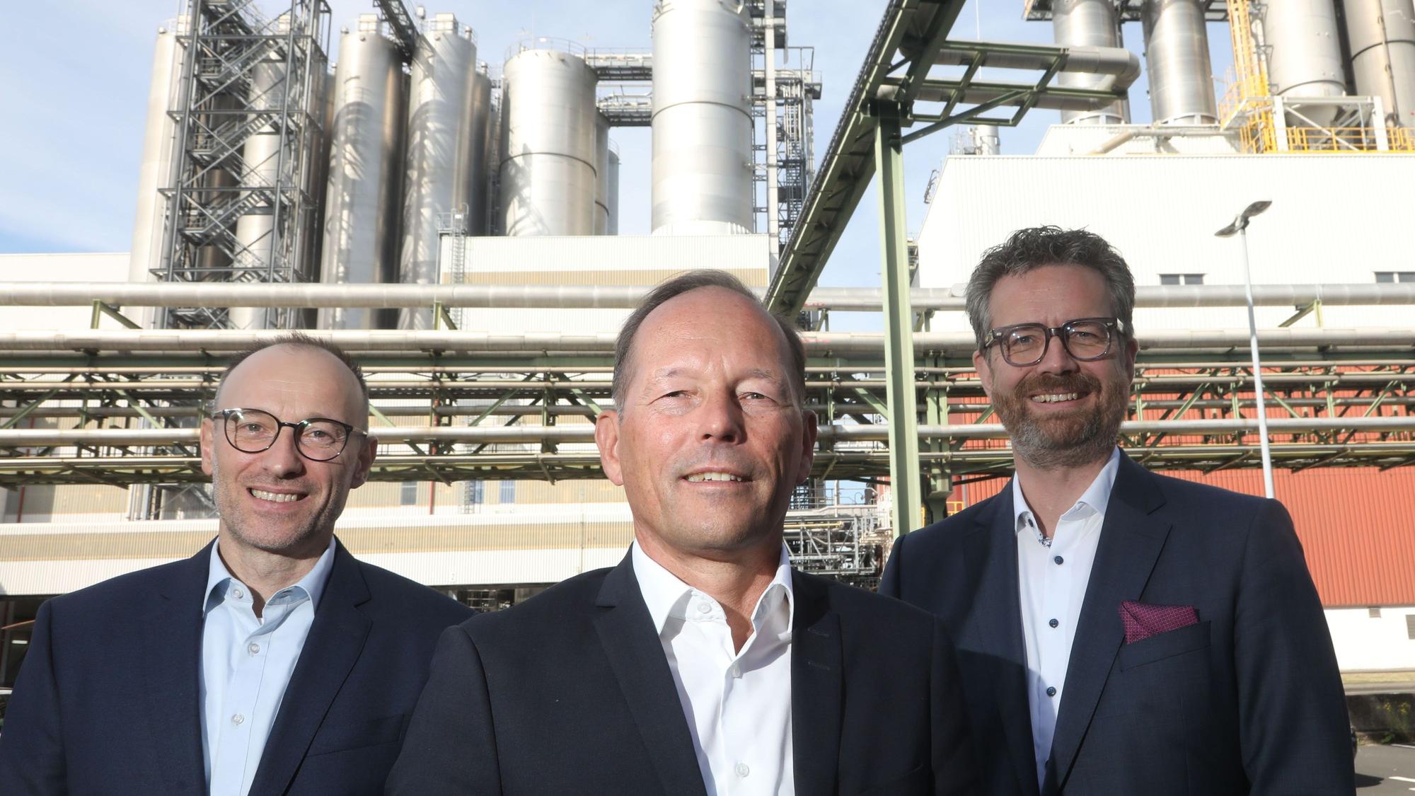 Gruppenbild mit Thomas Bastel, Thomas Wessel und Arndt Selbach. Alle tragen dunkle Anzüge ohne Krawatte. Im Hintergrund sind Chemieanlagen zu sehen.