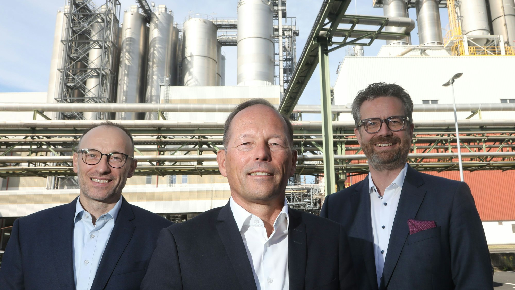 Gruppenbild mit Thomas Bastel, Thomas Wessel und Arndt Selbach. Alle tragen dunkle Anzüge ohne Krawatte. Im Hintergrund sind Chemieanlagen zu sehen.