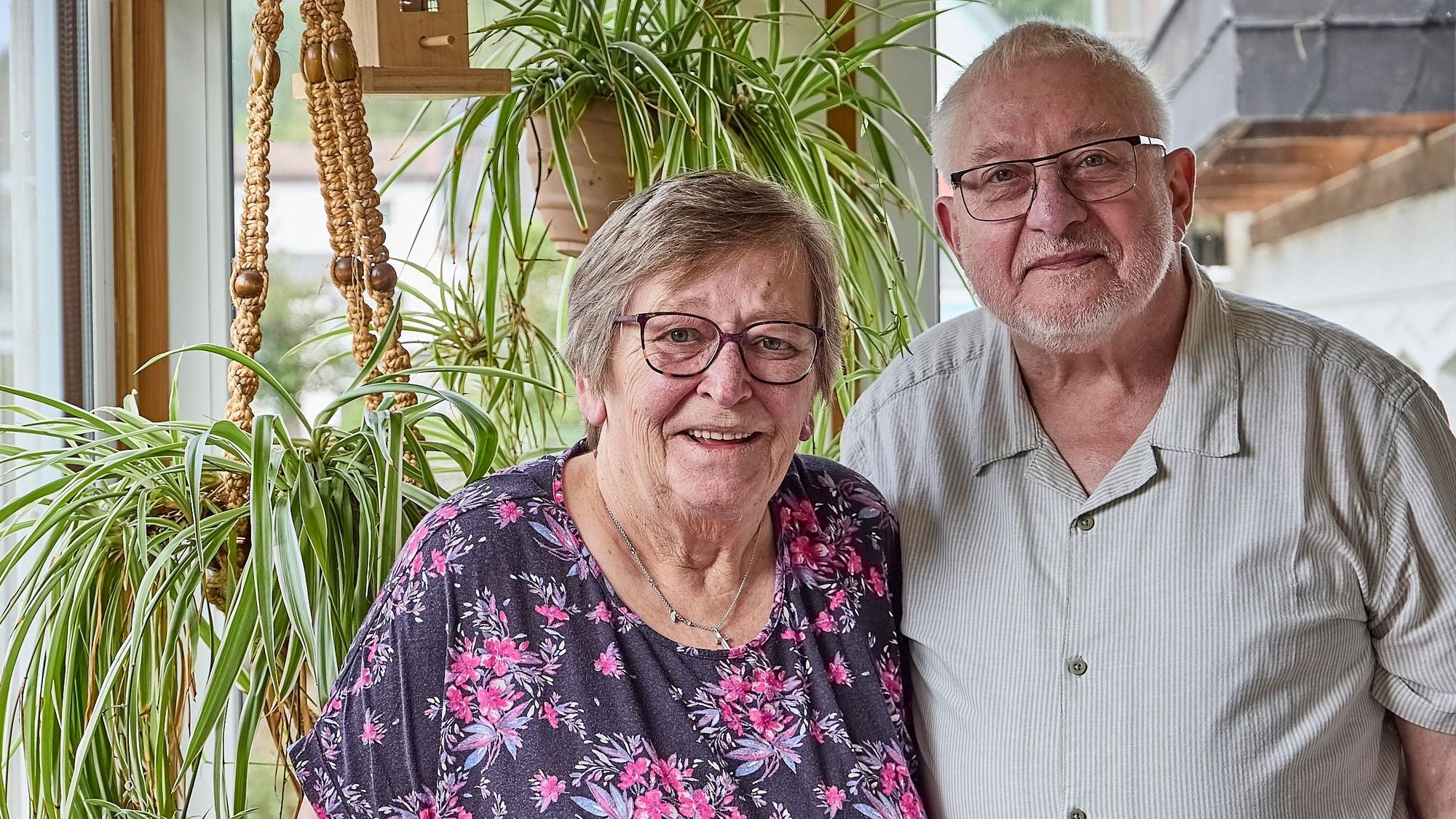 Betty und Hans Herzet stehen nah nebeneinander in einer Wohnung. Sie tragen beide Brille und lächeln in die Kamera. Im Hintergrund befinden sich Pflanzen.
