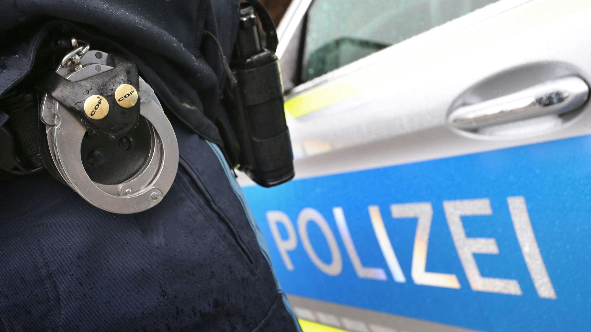 Ein Polizist mit Handschellen und einer Pistole am Gürtel steht vor einem Streifenwagen.