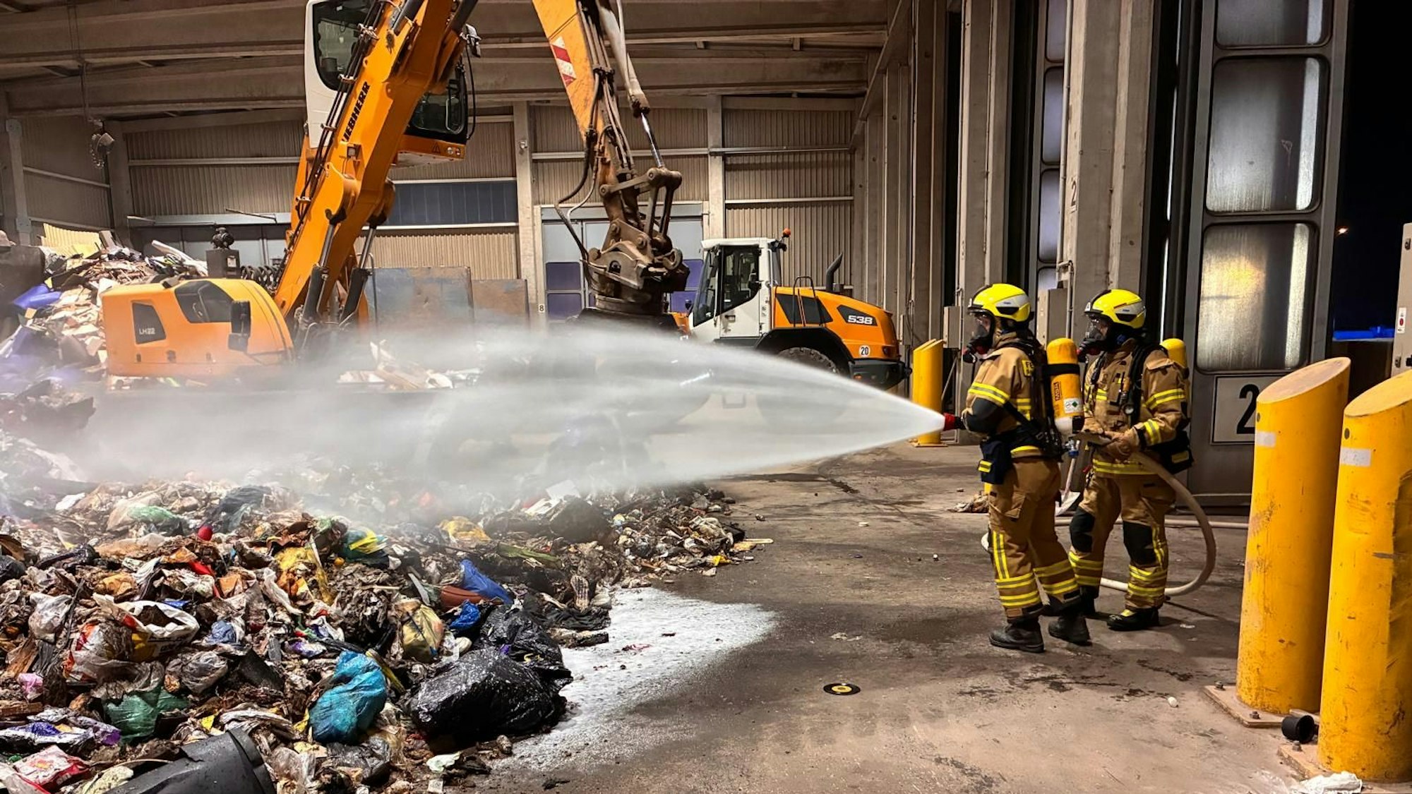 Zwei Feuerwehrleute stehen mit einem Löschschlauch vor einem Abfallhaufen, im Hintergrund ein Bagger.