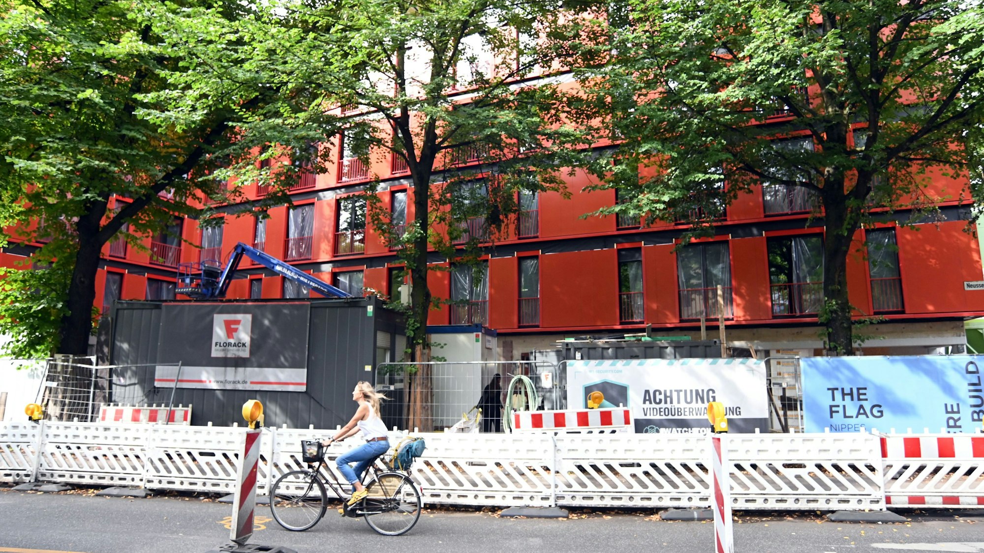 An einer Straße steht ein langgezogenes rotes Gebäude hinter einem Bauzaun.