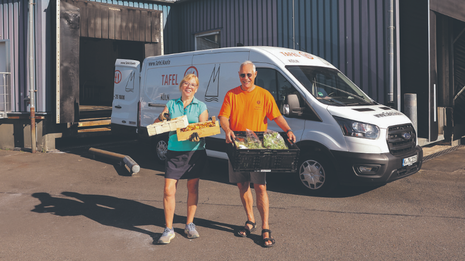 Ein Mann und eine Frau stehen vor einem weißen Ford Transit und halten Kisten mit Lebensmitteln in den Händen.