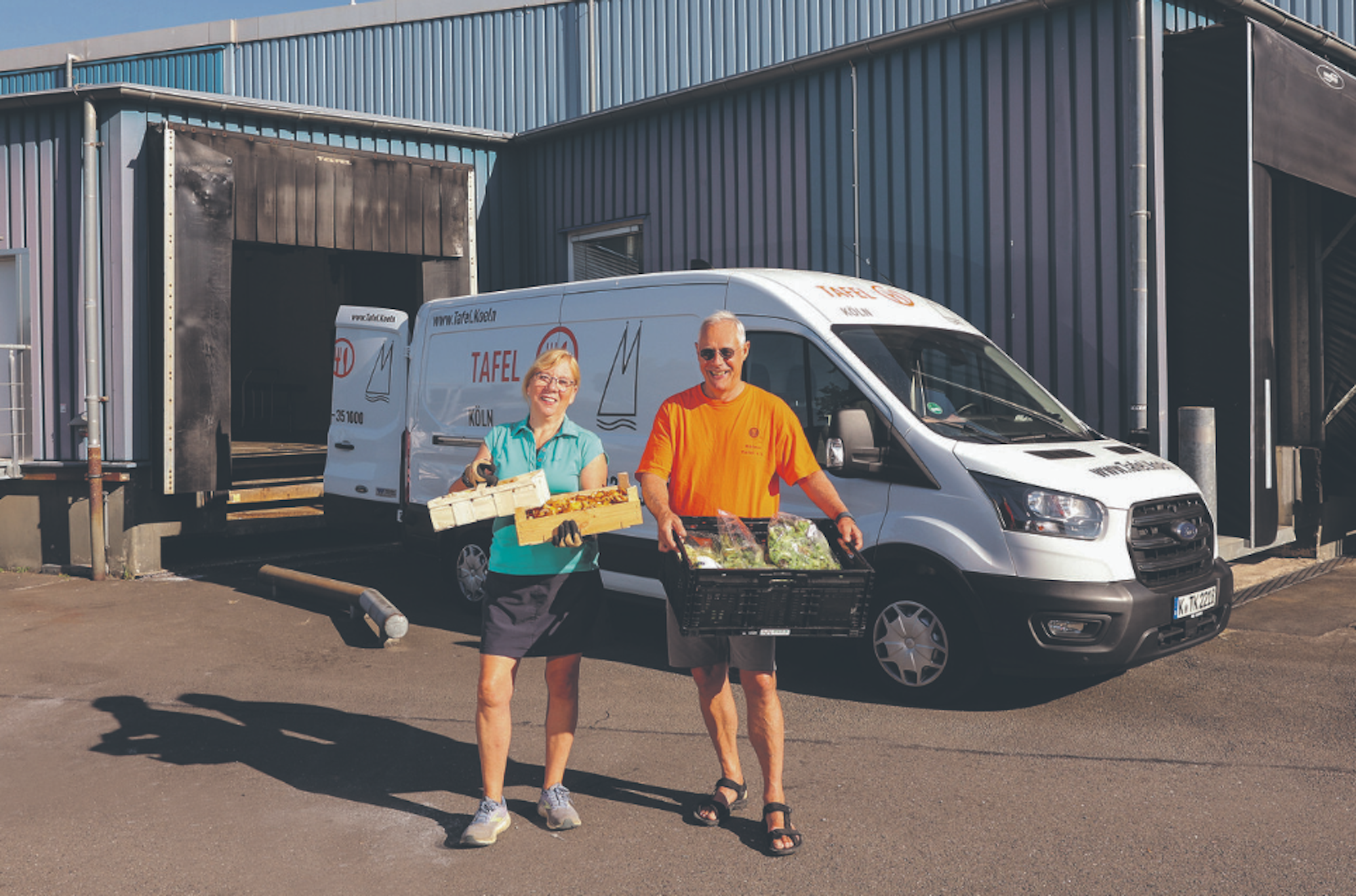 Ein Mann und eine Frau stehen vor einem weißen Ford Transit und halten Kisten mit Lebensmitteln in den Händen.