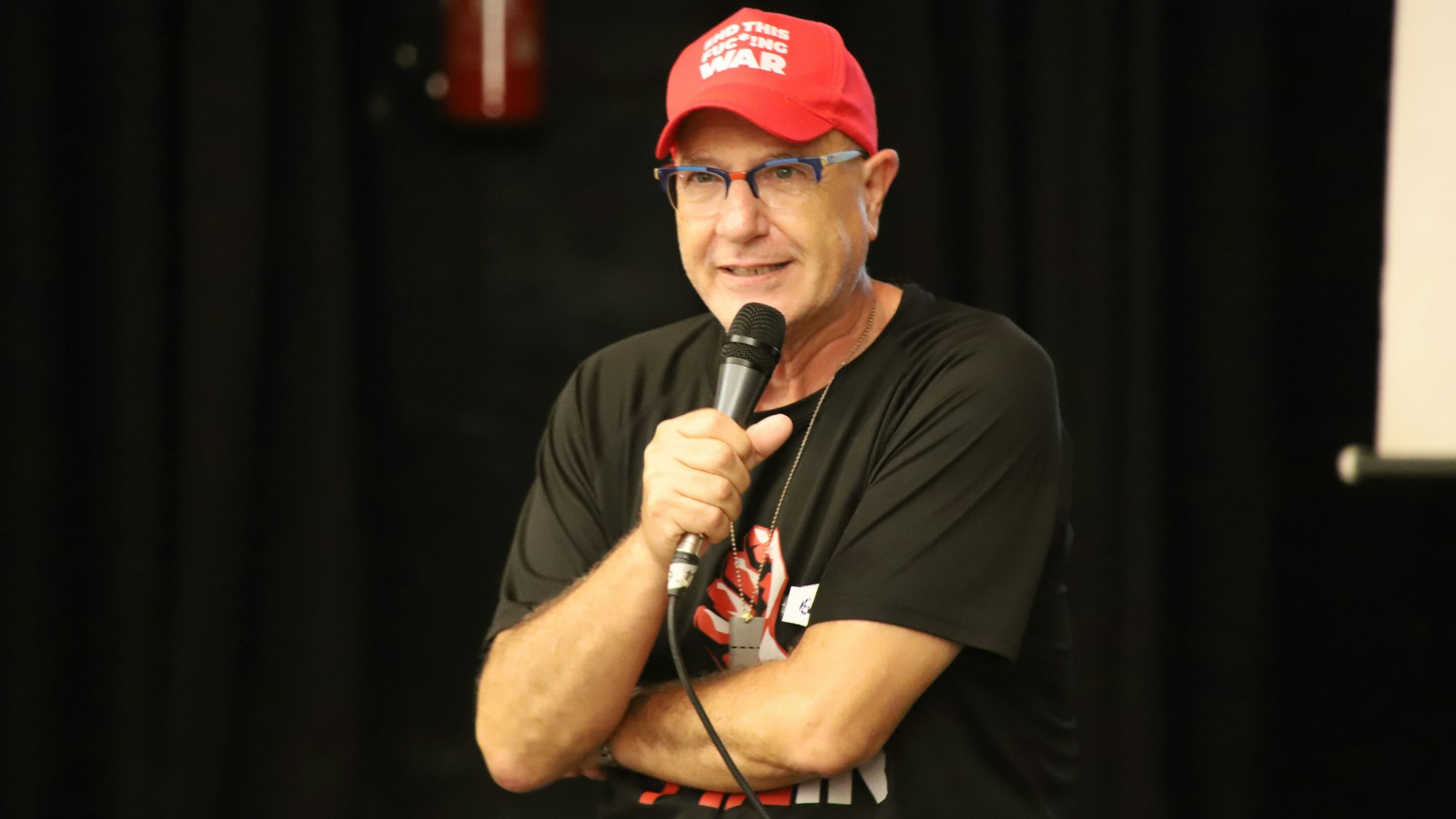 Ein Mann im schwarzen T-Shirt und mit einer roten Baseball-Kappe spricht in ein Mikrofon.