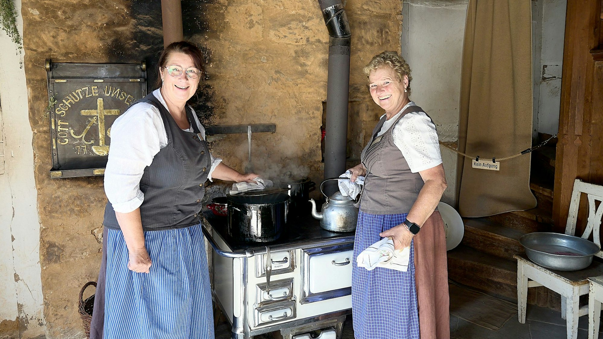 Zwei Frauen, Petra Spürkel und Anita Wolfgarten, stehen im Freilichtmuseum Kommern am Holzherd im Hof aus Elsig. Sie tragen traditionelle Kleidung mit blauen Schürzen.