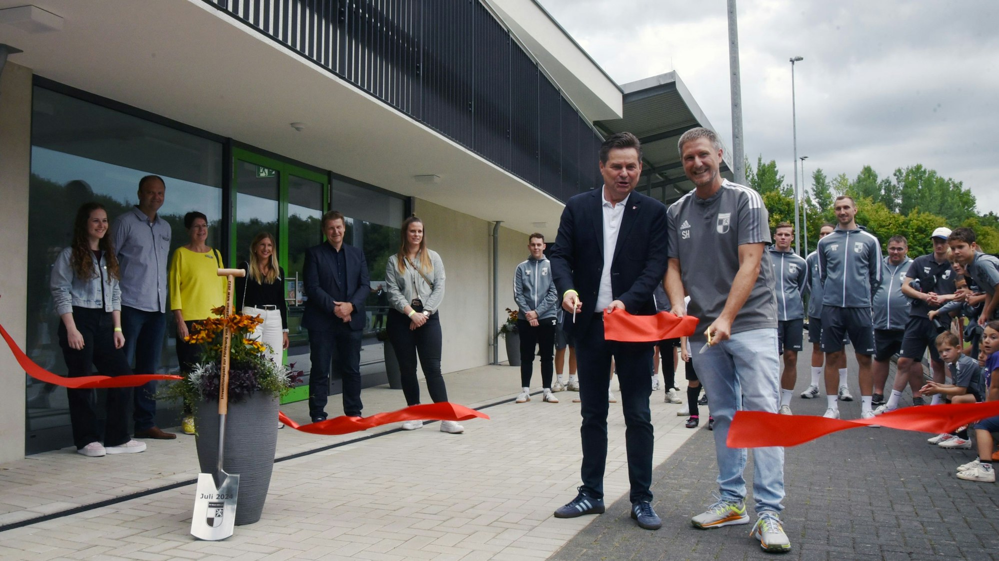 Zwei Männer mit Scheren in der Hand vor einem großen Zweckbau.
