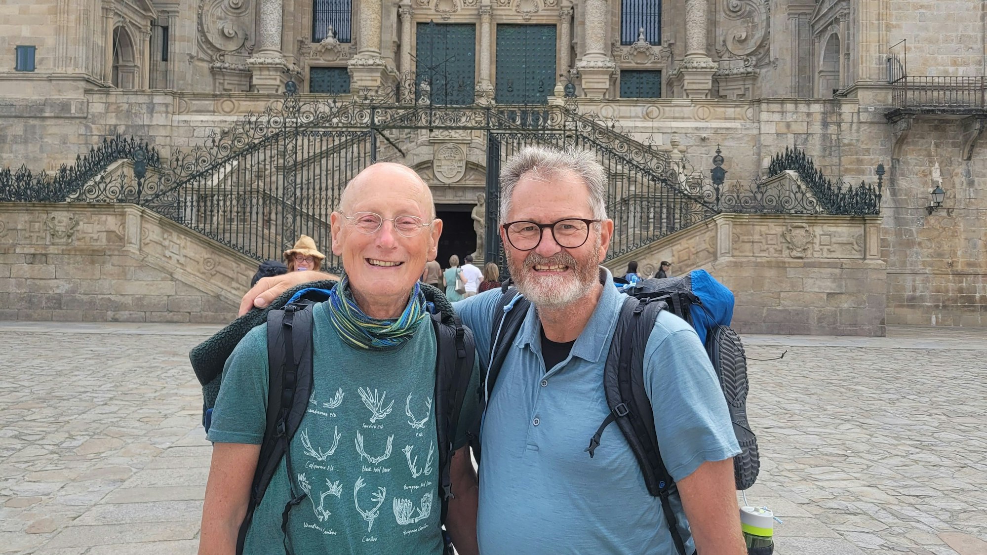 Zwei Männer mit Wandergepäck stehen Arm in Arm vor einer Kirche.