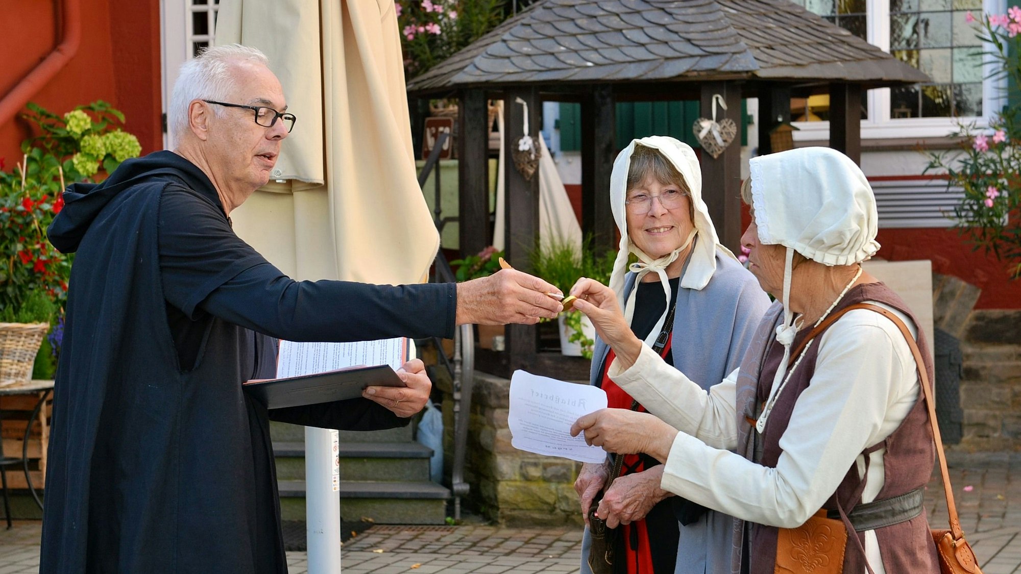 Ein Schauspieler, verkleidet als Pfarrer, bekommt von zwei weiteren Schauspielerinnen Goldmünzen überreicht.