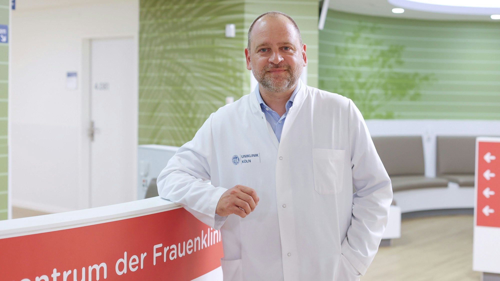 Herr Taran
ist im Halbporträt und Arztkittel zu sehen im Eingangsbereich der Kölner Frauenklinik.