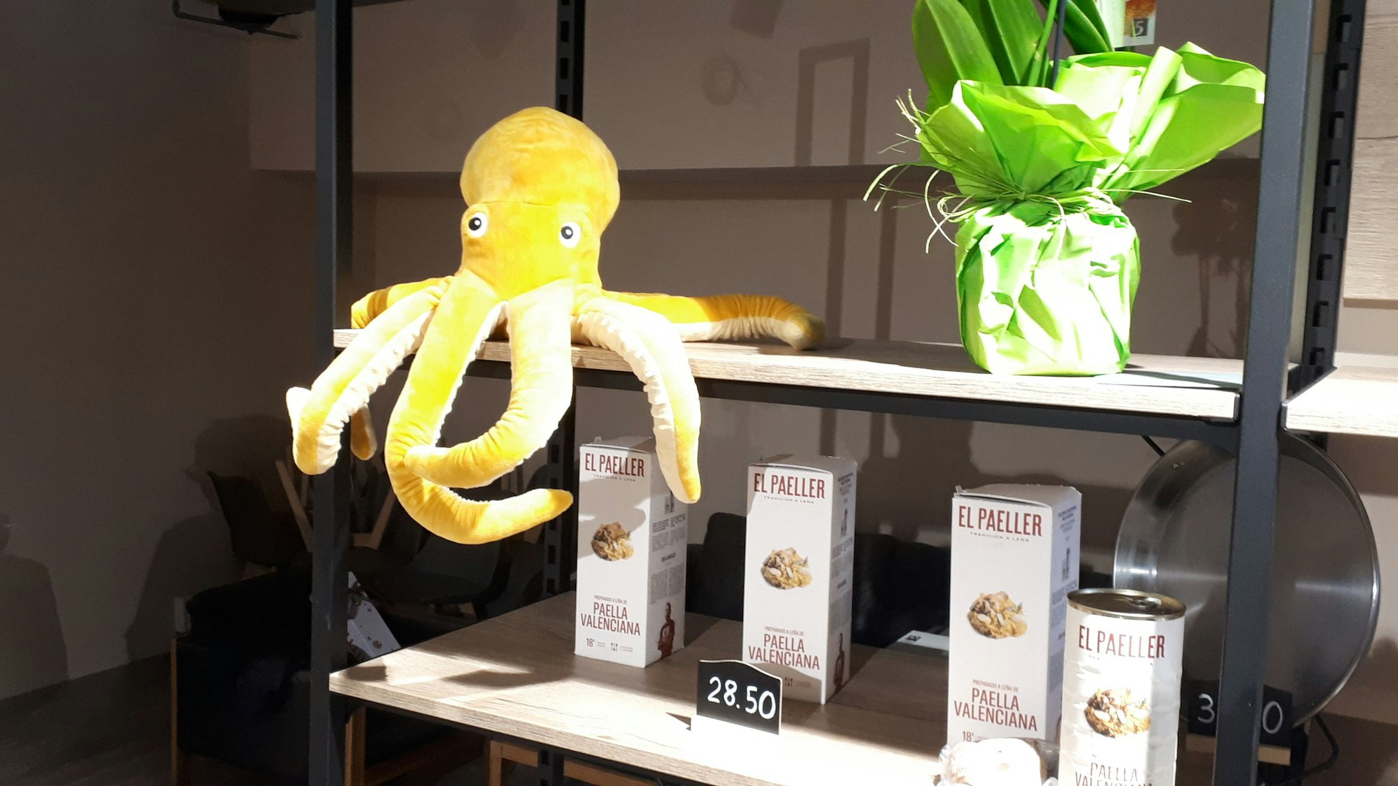 Ein gelber Plüsch-Oktopus sitzt auf einem Regal, in dem verschiedene Paella-Pakete stehen.