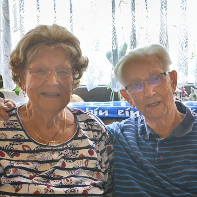 Gertrud Elisabeth und Rudolf Pohl sitzen vor einem hellen Vorhang, durch den Licht scheint. Er hält seinen rechten Arm um sie.