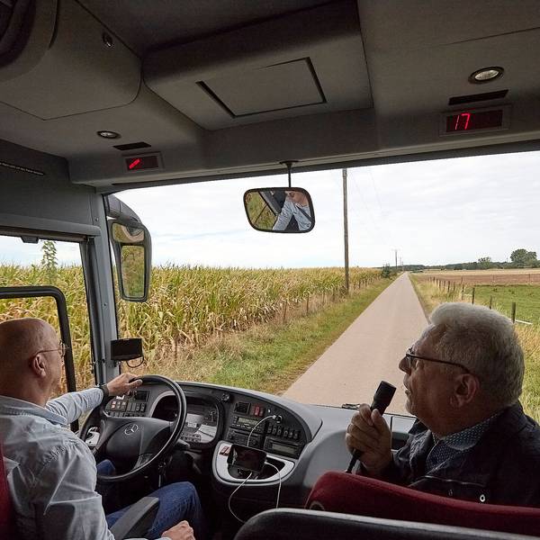 Ein Bus fährt über einen Wirtschaftsweg an einem Maisfeld entlang. Neben dem Fahrer sitzt der Bürvenicher Ortsvorsteher Jörg Körtgen und spricht in ein Mikrofon.