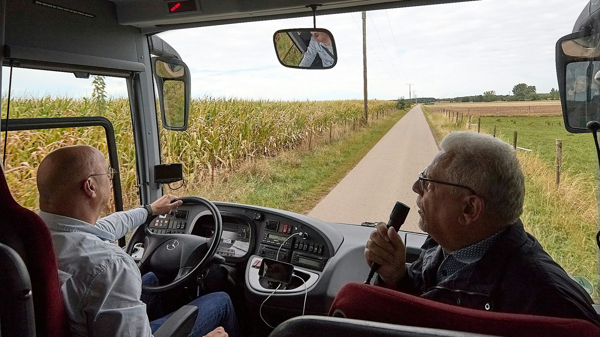 Ein Bus fährt über einen Wirtschaftsweg an einem Maisfeld entlang. Neben dem Fahrer sitzt der Bürvenicher Ortsvorsteher Jörg Körtgen und spricht in ein Mikrofon.