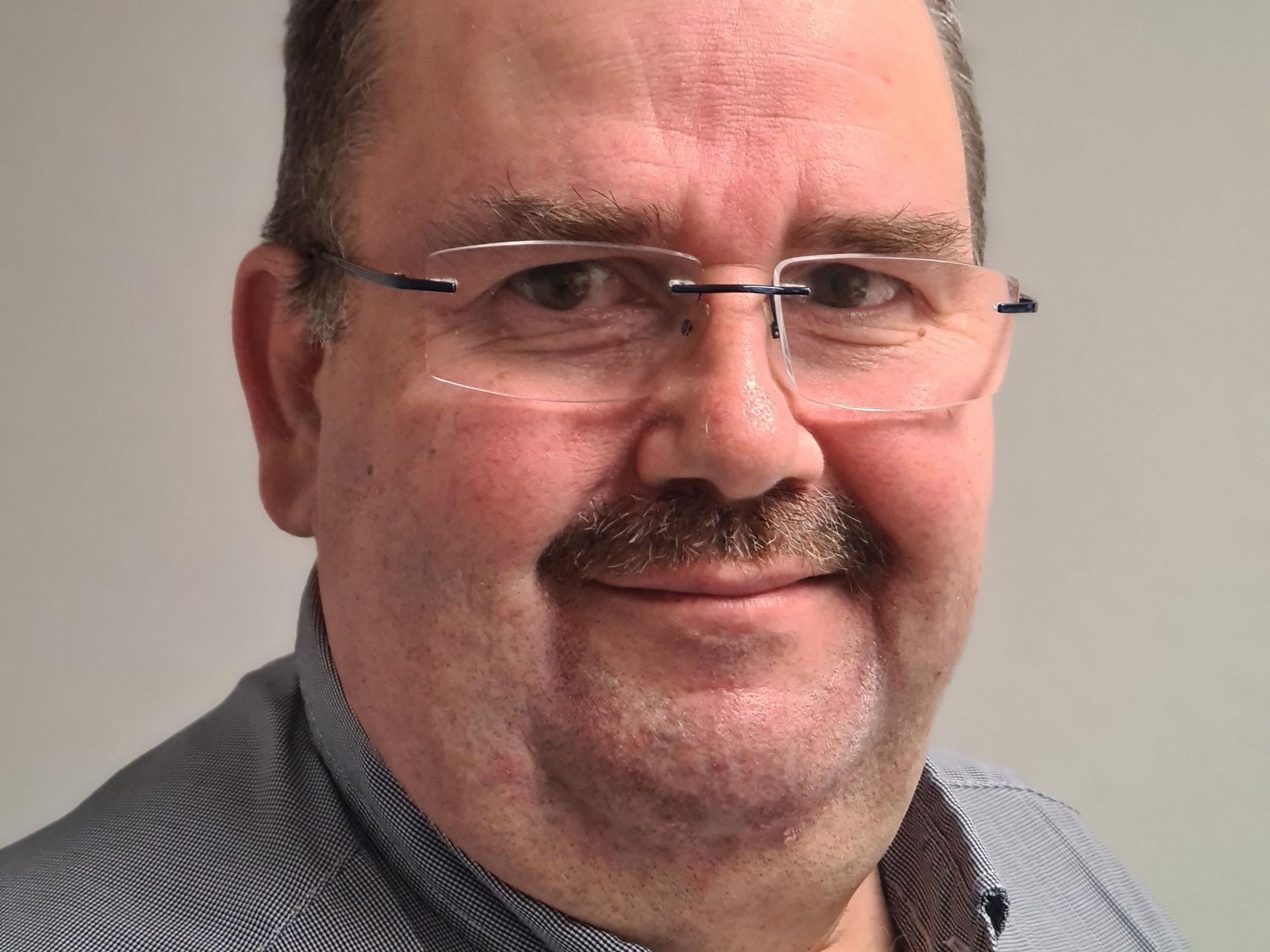 Linke trägt ein graues Hemd und eine Brille.