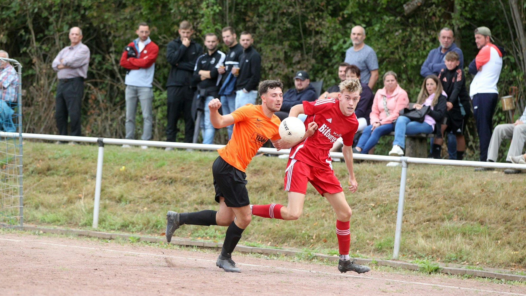 Zwei Fußballer kämpfen auf einem Aschenplatz an der Seitenlinie um den Ball. Im Hintergrund beobachten die Zuschauer die Szene.