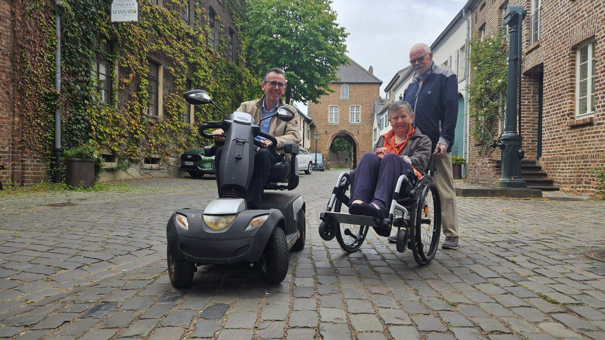 Zu sehen sind ein Mann in einem E-Mobil und ein Mann, der eine Frau im Rollstuhl über ein Kopfsteinpflaster schiebt.