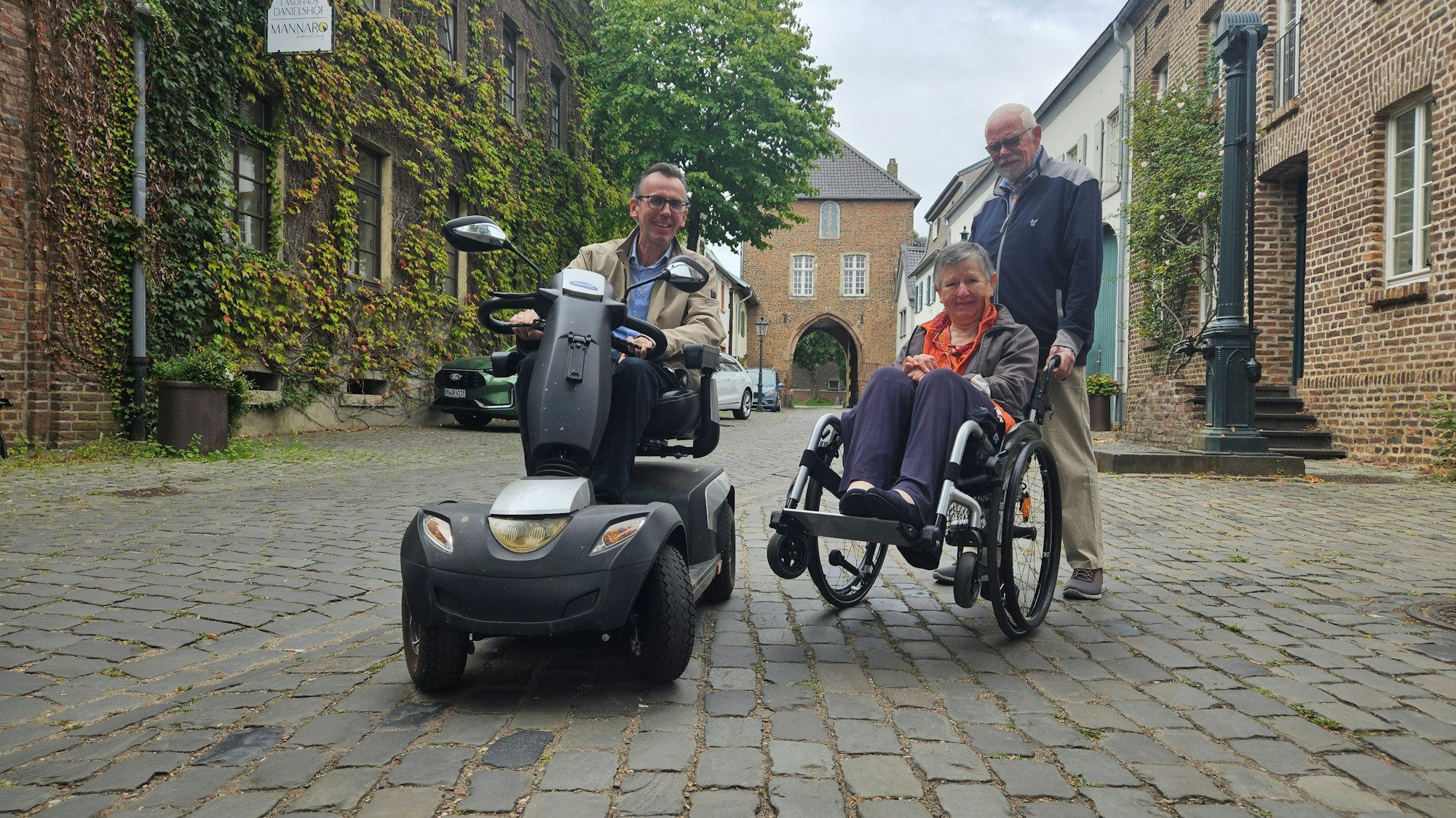 Zu sehen sind ein Mann in einem E-Mobil und ein Mann, der eine Frau im Rollstuhl über ein Kopfsteinpflaster schiebt.