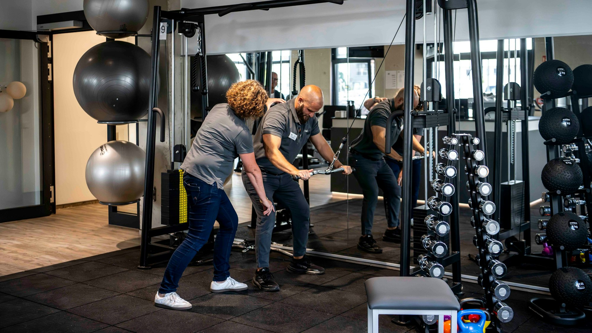 Das Bild zeigt das Fitness- und Kraftstudio der Physiotherapie-Spezialisten. Ein Mann macht eine Sportübung und wird dabei angeleitet.