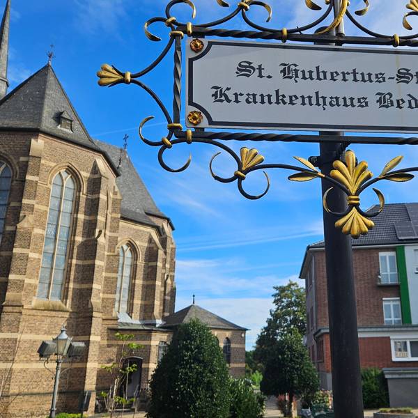 Zu sehen ist das Bedburger Krankenhaus neben der Kirche, im Vordergrund steht ein Schild, das auf das Krankenhaus verweist.
