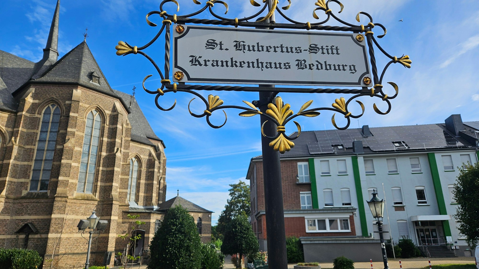 Zu sehen ist das Bedburger Krankenhaus neben der Kirche, im Vordergrund steht ein Schild, das auf das Krankenhaus verweist.