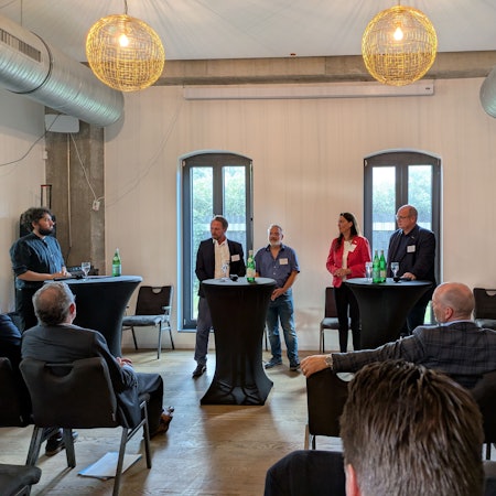 Sechs Männer und eine Frau stehen an Stehtischen und debattieren vor Publikum.