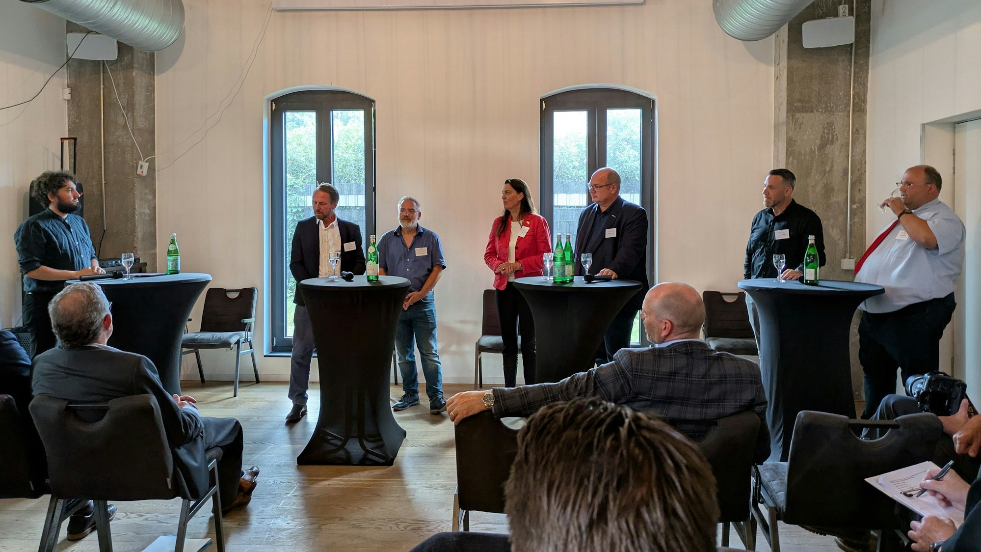 Sechs Männer und eine Frau stehen an Stehtischen und debattieren vor Publikum.