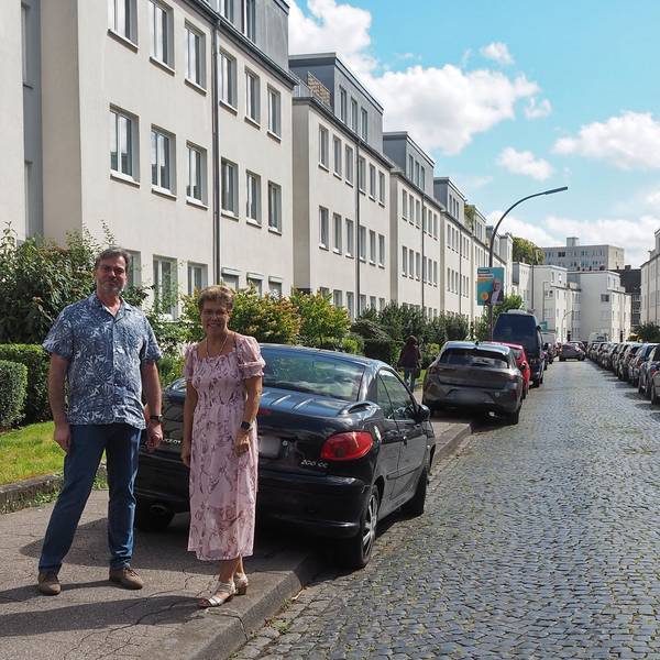 Ein Mann und eine Frau stehen auf einem Bürgersteig, an dessen Straße viele Autos geparkt sind.