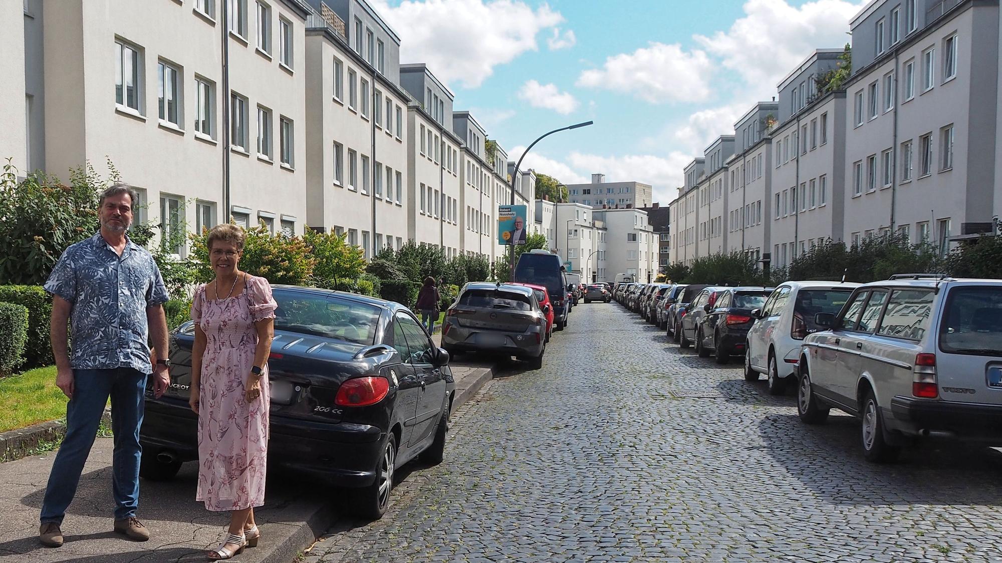 Ein Mann und eine Frau stehen auf einem Bürgersteig, an dessen Straße viele Autos geparkt sind.