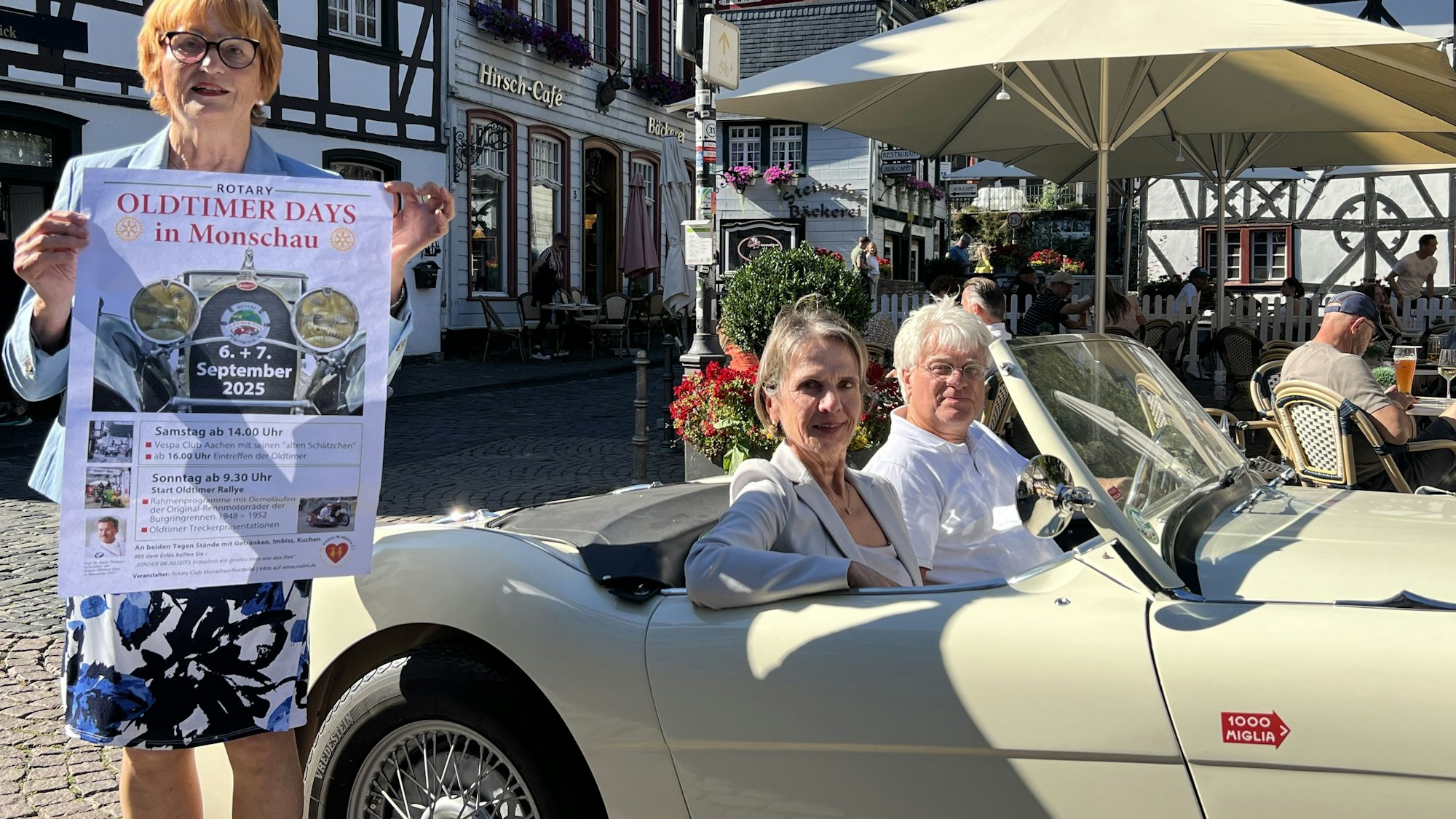 Eine Frau steht auf dem Marktplatz in Monschau und hält ein Plakat in den Händen. Daneben sitzen ein Mann und eine Frau in einem Austin-Healey-Oldtimer.