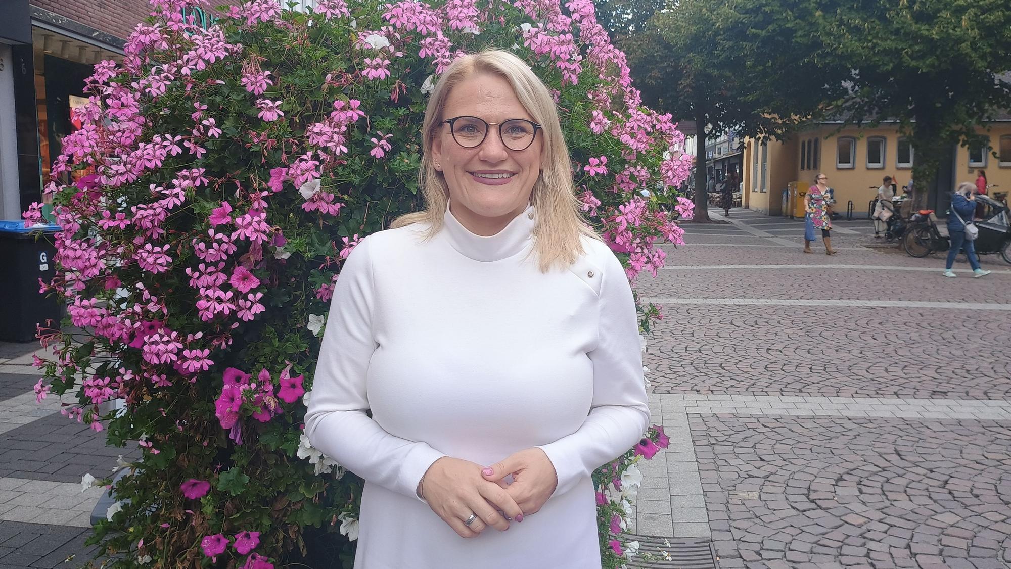 Christina Stumpp steht in der Brühler Innenstadt vor einem blumigen Busch. Sie trägt ein weißes Oberteil und eine Brille.