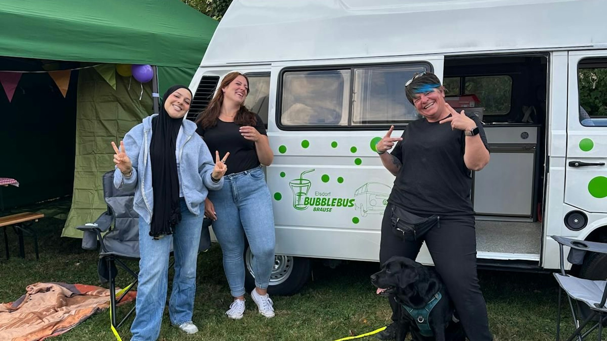 Das Bild zeigt die drei Mitarbeitende vor dem weißen Bus, auf dem grüne Punkte zu sehen sind.