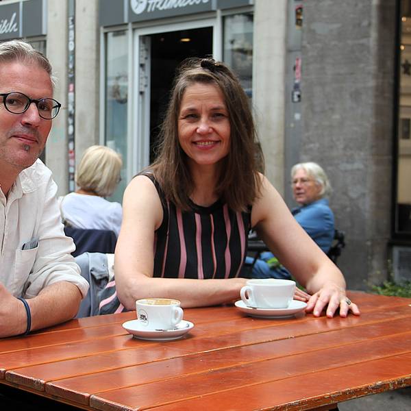 Ein Mann und eine Frau sitzen draußen in einem Café an einem Holztisch, auf dem zwei Kaffeetassen stehen.
