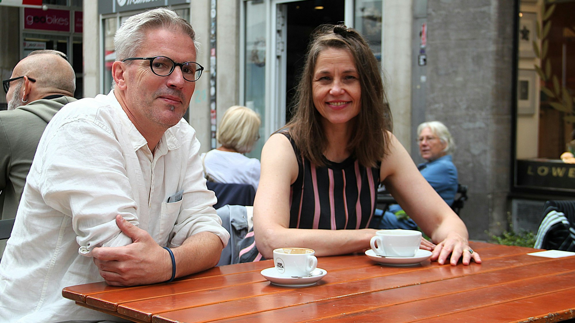Ein Mann und eine Frau sitzen draußen in einem Café an einem Holztisch, auf dem zwei Kaffeetassen stehen.