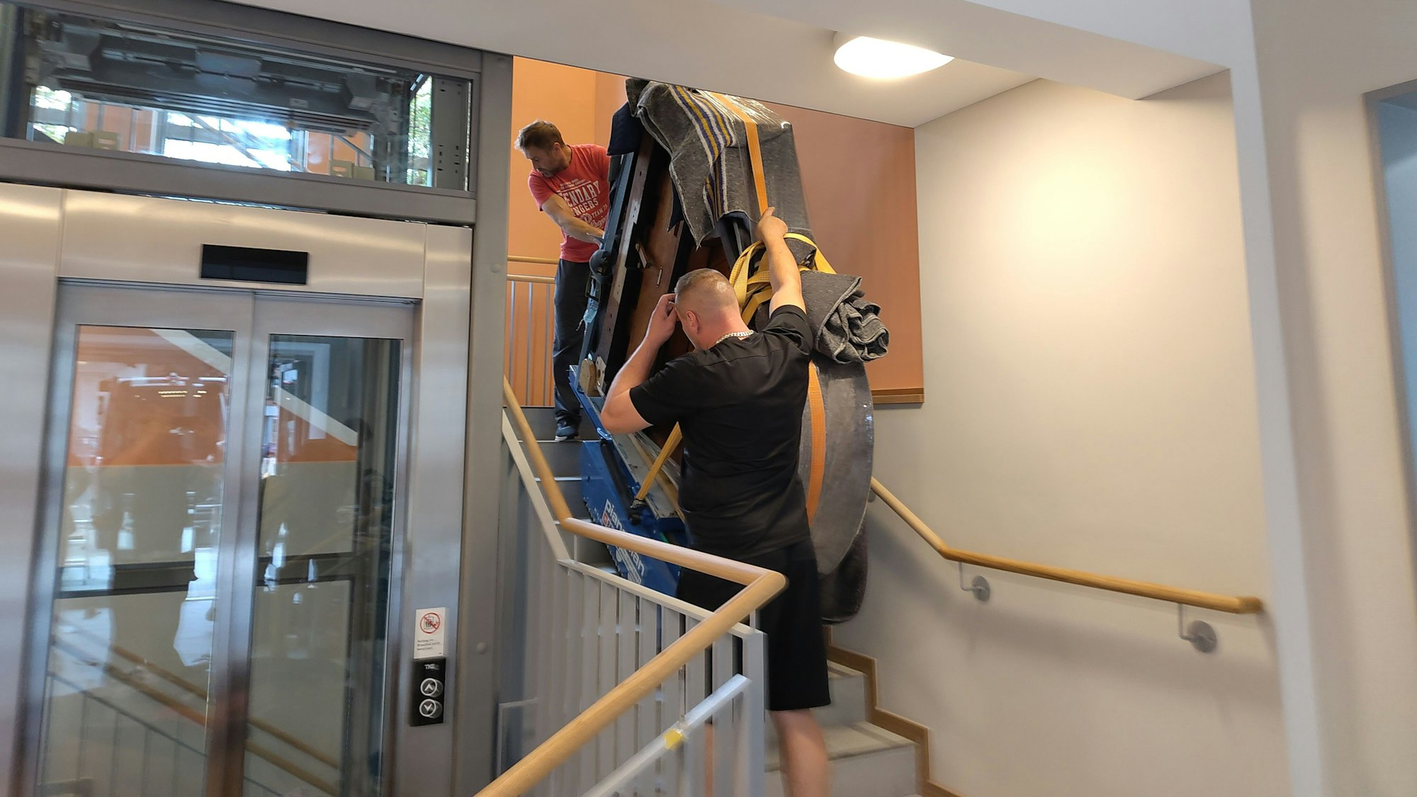 Möbelpacker tragen einen Flügel eine Treppe hinauf.