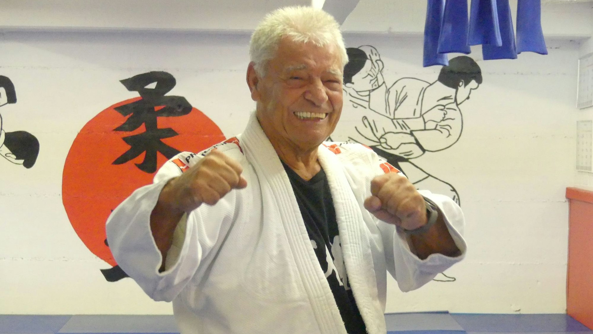 Ein Mann im Judo-Dress steht in einer Halle, die mit japanischen Zeichen und Bildern vom Judo geschmückt ist.