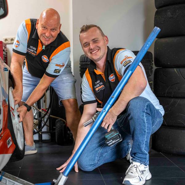 Reiner und Yannick Habeth grinsen neben einem Rennauto. Reiner Habeth fasst an den vorderen Reifen des Fahrzeugs. Yannick Habeth kniet daneben und hält Werkzeug.