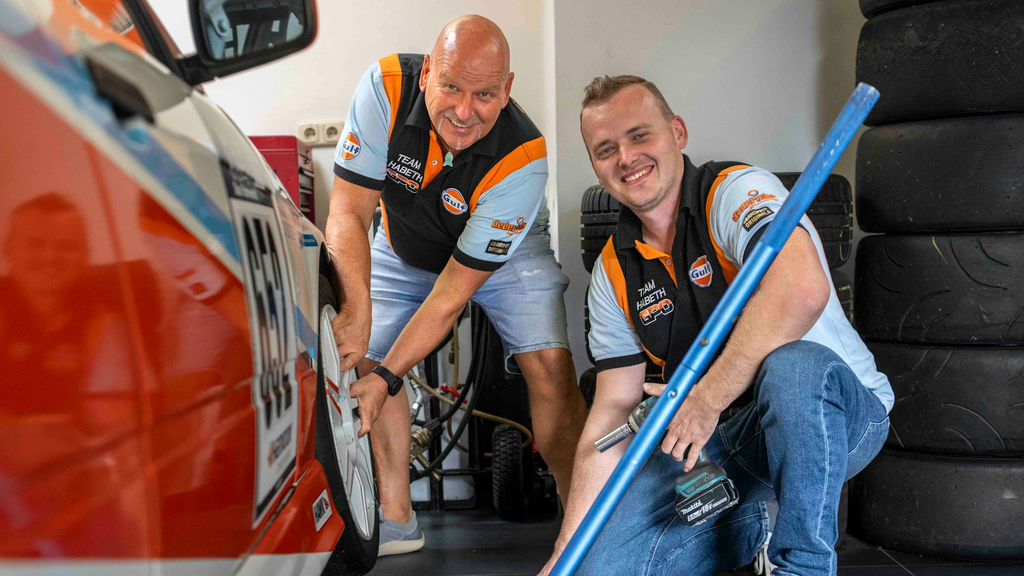 Reiner und Yannick Habeth grinsen neben einem Rennauto. Reiner Habeth fasst an den vorderen Reifen des Fahrzeugs. Yannick Habeth kniet daneben und hält Werkzeug.