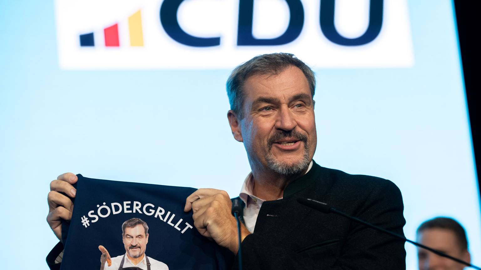 Markus Söder (CSU) steht auf der Bühne und zeigt eine Grillschürze mit der Aufschrift „#SÖDERGRILLT“.