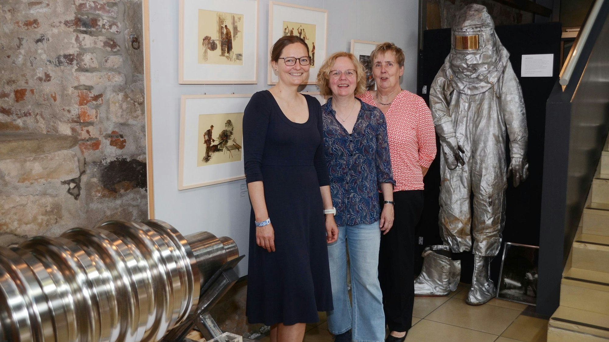 Drei Frauen in einer Ausstellung. Vorne liegt ein schweres, auf Hochglanz poliertes Maschinenteil, im Hintergrund ist ein Sicherheitsanzug für die Arbeit am Hochofen zu sehen.