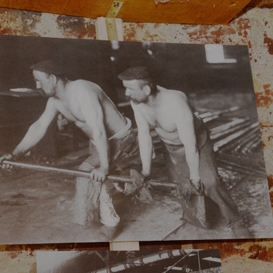 Eine historische Aufnahme zeigt Männer mit freiem Oberkörper bei der Arbeit in einer Fabrik.