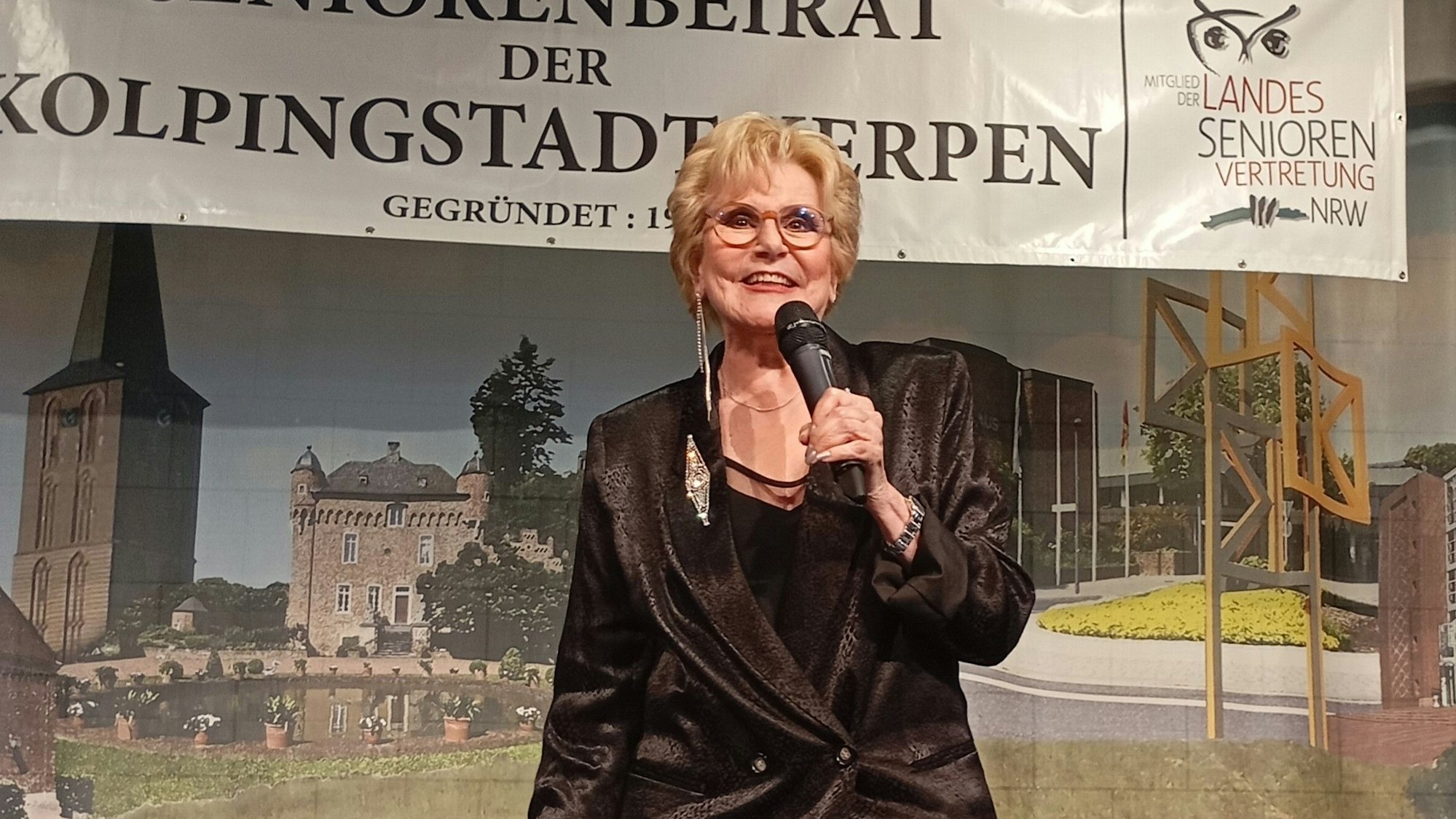 Peggy March steht mit einem Mikrofon auf der Bühne, sie hat kurze blonde Haare und trägt ein schwarzes Sakko.