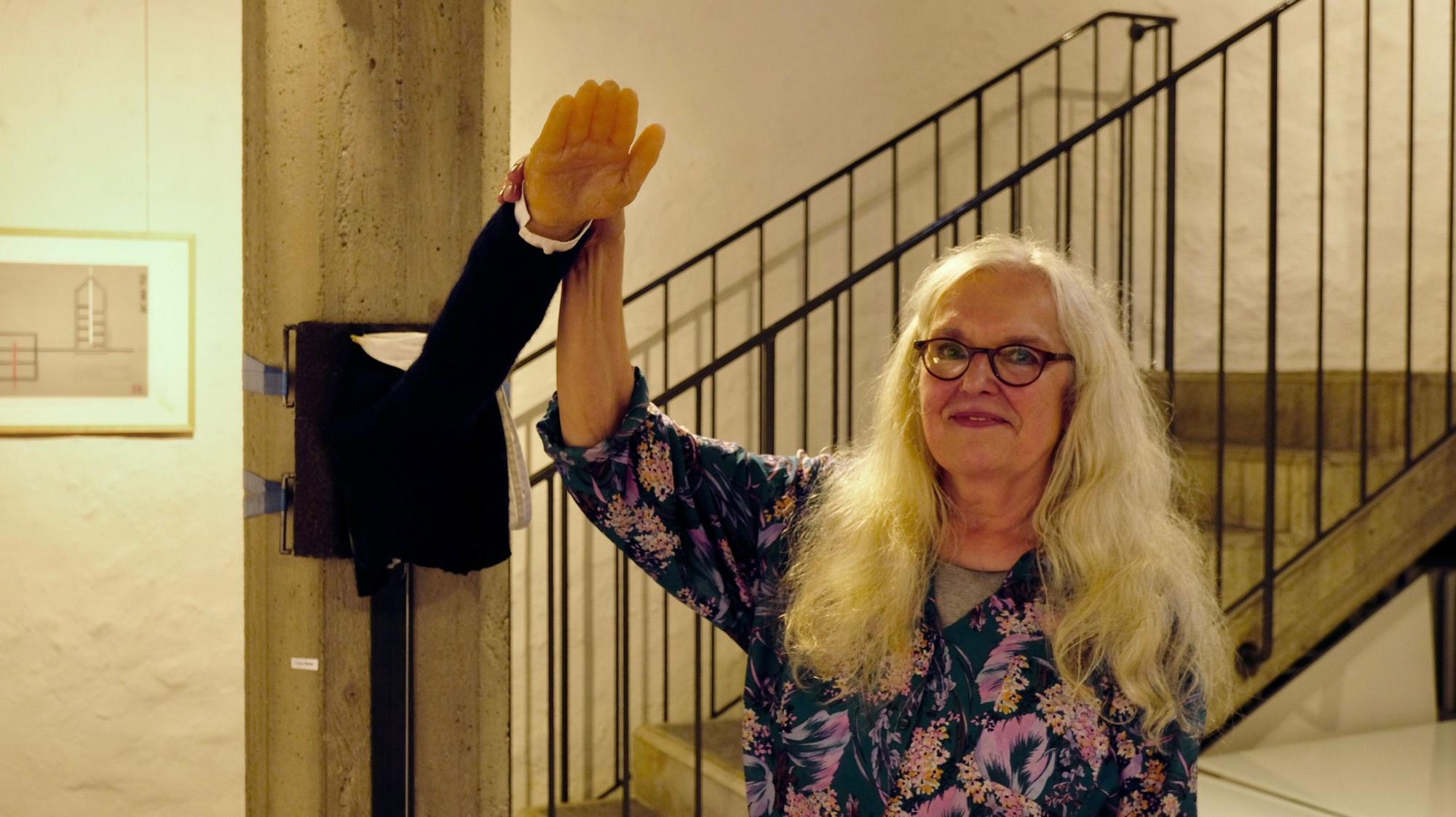 Eine Frau mit langem blonden Haar und Brille hält einen aus der Wand ragenden Arm, bei dem es sich um eine Kunstinstallation mit dem Titel "Tu was" handelt.