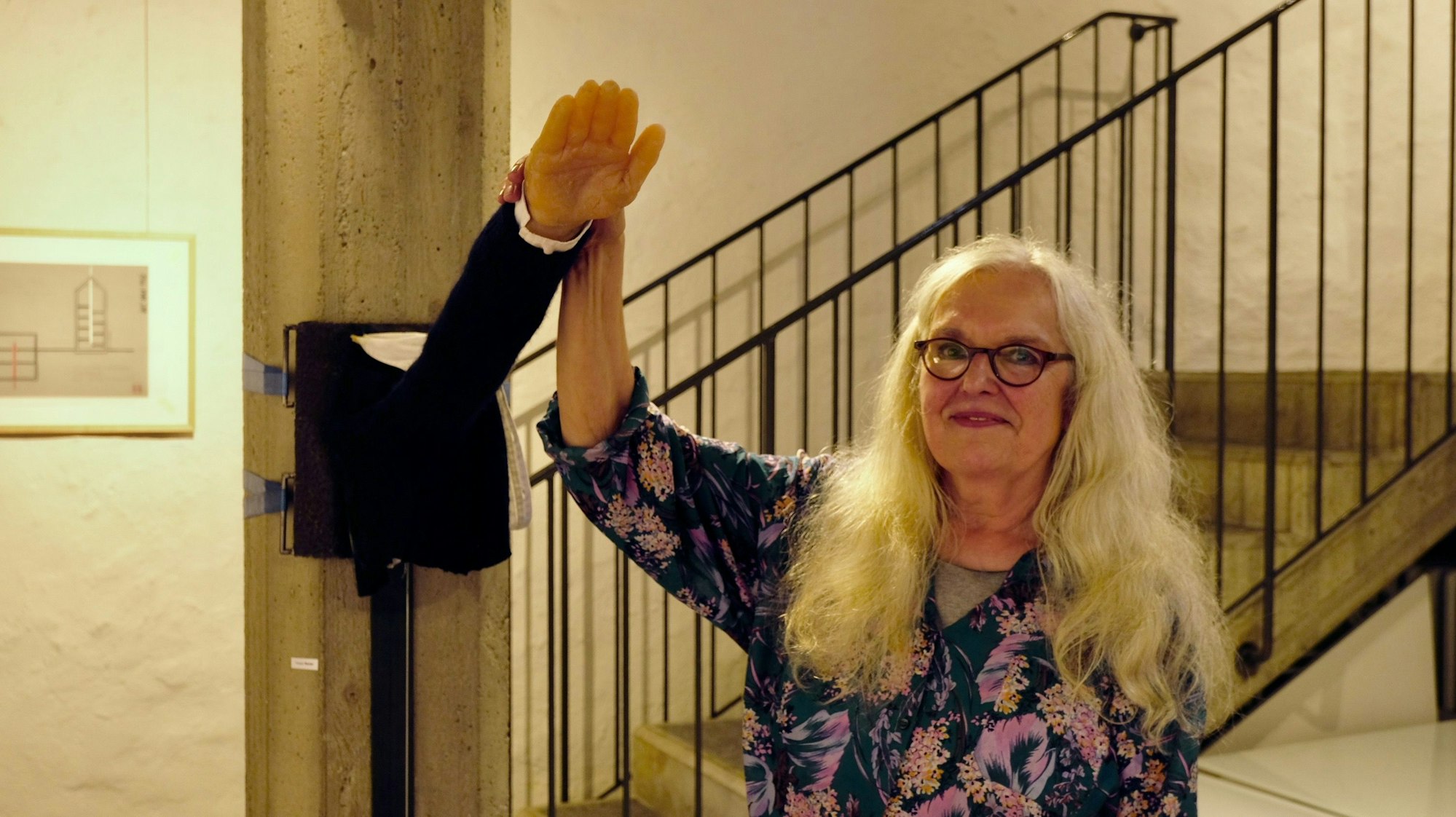Eine Frau mit langem blonden Haar und Brille hält einen aus der Wand ragenden Arm, bei dem es sich um eine Kunstinstallation mit dem Titel "Tu was" handelt.