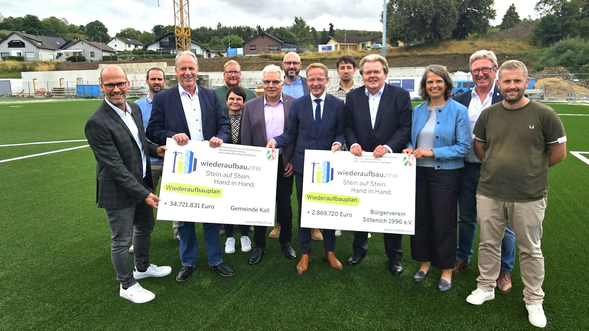 Eine Gruppe Menschen steht auf dem Kunstrasenplatz in Sötenich. Sie halten zwei Förderbescheide für die Gemeinde Kall und den Bürgerverein in den Händen.